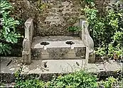 A stone bench-like construction with two holes in it, in front of a brick wall. There are plants growing around it.
