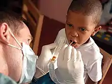 white man holding plastic tray with brown goop in it and sticking a small stick into a black boy's open mouth
