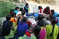 A woman teaches women's literacy class. There is a group of women surrounding a single woman who holds up a board on which there is foreign writing. She is teaching literacy to the group of women.