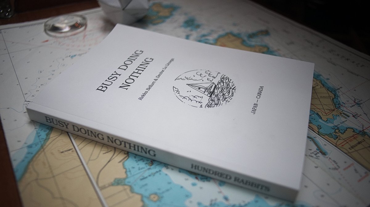 A book with a white cover laying over a chart, the title reads Busy Doing Nothing and features a small illustration of a boat riding waves that look like rabbits