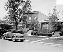 Two-story house with cars parked on the street in front of it. It is partially obstructed by a neighboring house on the right. A tall tree is on the lawn in front of the house.