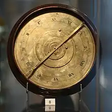 A brass, clock-like mechanical device in a museum display case, with a small card with the number 8 printed on it. The face of the device is split into several rings, with the Roman numerals I through XI (and 0) on one of these rings.