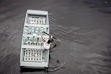 Overhead view of a young girl, alone, rowing through murky floodwaters on a wooden boat carrying several large plastic water bottles