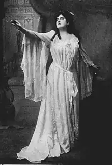 A white woman in a flowing gown, standing with one arm outstretched.