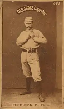 A baseball-card photograph of a mustachioed man in an old-style white baseball uniform holding a baseball in front of his chest with both hands