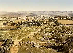 A colorized skyline of a town with  hills on the horizon, white buildings in the background, and a cemetery, olive trees, and dirt paths in the foreground
