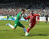 A soccer player dressed in green and white runs with the ball. A player in red and white tries to take the ball. In the background there are stands filled with Hammarby fans.