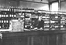 A photo of a man behind the counter at a liquor store with shelves stocked with liquor behind him.