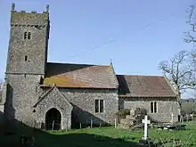 A stone church seen from the south, with a square battlemented tower at the left, then the nave, and a lower chancel at the right. In front of the church is the base of a medieval cross