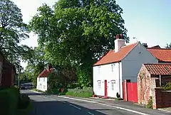 A pair of whitewashed cottages, with pantiled roofs, widely spaced along a road. the nearer one has a red gate and a small pantiled outhouse. A huge oak, in full leaf, stands behind the further one.