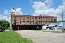 The same brick building seen from a different angle. An American flag flies from a short tower at the left end, and a flat roof projects over part of a parking area on that side at ground level. A one-story white building is at the right.