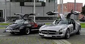 Two cars with open gullwing doors: an older black one and a gray one