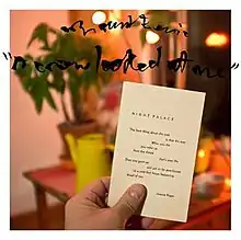 A man's hand holding the Joanne Kyger poem "Night Palace" in the foreground. Background is of a house illuminated by an orange light.