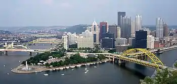 A view of a city nestled at the point where two rivers merge. There are yellow bridges crossing the rivers and a large fountain at the point where they meet. The city steps back from a park surrounding this fountain.