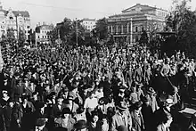 A crowd of civilians are standing in the foreground watching armed soldiers in the middle-ground marching alongside the street. Buildings of downtown Dresden can be seen in the background.