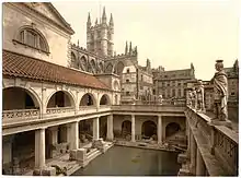 A late-nineteenth-century Photochrom of the Great Bath at the Roman Baths. Pillars tower over the water, and the spires of Bath Abbey – restored in the early sixteenth century – are visible in the background.