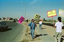 A male presenting person stands in the middle of the photograph holding two large signs on wooden sticks. The sign on the left is pink with black writing and reads "Stop Homophobia". The sign on the right is yellow with black writing and reads "We're here we're queer". The image is obviously old due to under exposure. There is another male presenting person on the right with his back to the camera holding similar signs. One which is yellow with a pink border and reads "Danne-Meyer = Death". The on on the right is cut off my the camera. Both individuals are standing on the side of a road way as a car drives by.