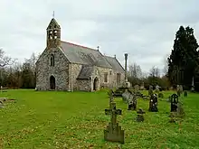 A stone church seen from the southwest, with a slate roof and red tiled ridge. At the west end is a door a window and a double bellcote; protruding from the south side is a porch