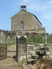 A small stone church standing in a churchyard on top of a hill. On the near gable is a bell-cupola.
