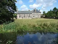 Seen beyond a river and a field is a simple stone chapel with three arched windows and a door; to the right is a two-storeyed stone house