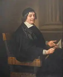 A man with a black cap and gown, seated. He is holding an open book in his right hand and pointing to it with his left