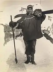 Man, standing, wearing a smock, heavy trousers and boots. He has a ski stick in his right hand, a pair of skis strapped on his back, and is carrying a rounded bundle on his shoulder. Behind him on the ground is assorted polar equipment.