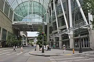 A street under a tall, arched glass bridge, as seen from a sidewalk