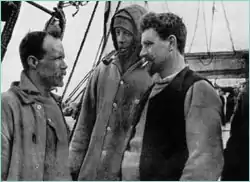 Three men in well-worn clothing stand on the deck of a ship. Man on right is thickset, bearded, with cigarette in mouth. Man in centre is hooded; man on left, also bearded and with receding hair, is shown right profile.