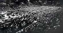Thousands of spectators in a large arena look towards a man standing behind a podium on a platform